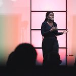 Dagvoorzitter met microfoon op een podium voor een volle zaal